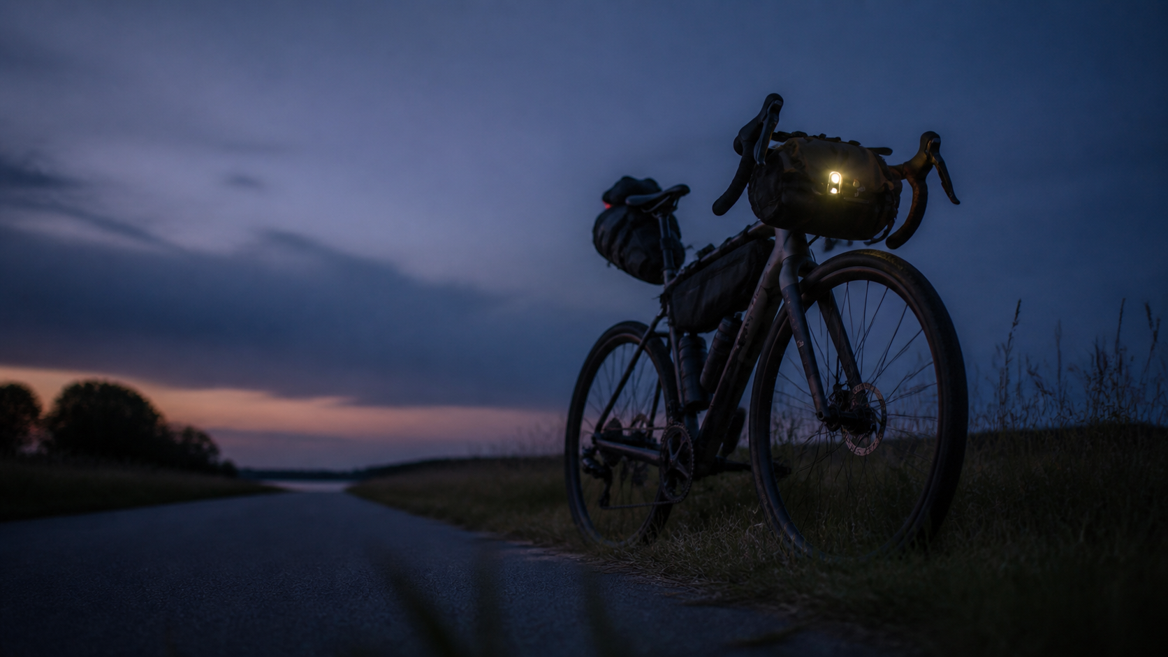 A bikepacking-rigged gravel bike with a glowing front light stands beside an empty road at dusk, warm light on the horizon.